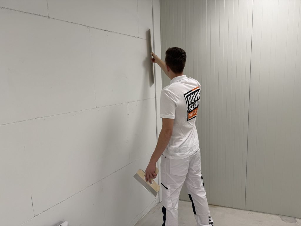 Stukadoor in wit werkpak trekt met een lange spaan een wand van gipsblokken vlak, plamuurmes in de andere hand